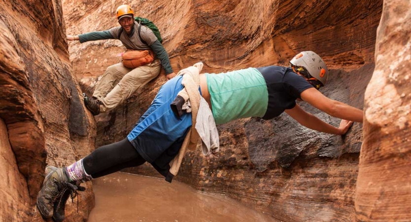 A person wearing a helmet braces themselves on either side of a slot canyon. There is water below them. 